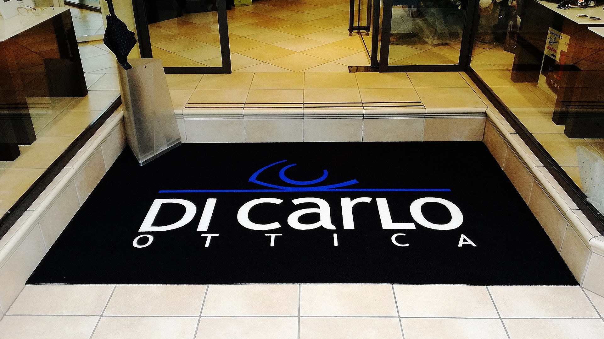 Ingresso di un negozio con un Tappeto intarsiato personalizzato con il logo Di Carlo Ottica di colore nero, blu e bianco